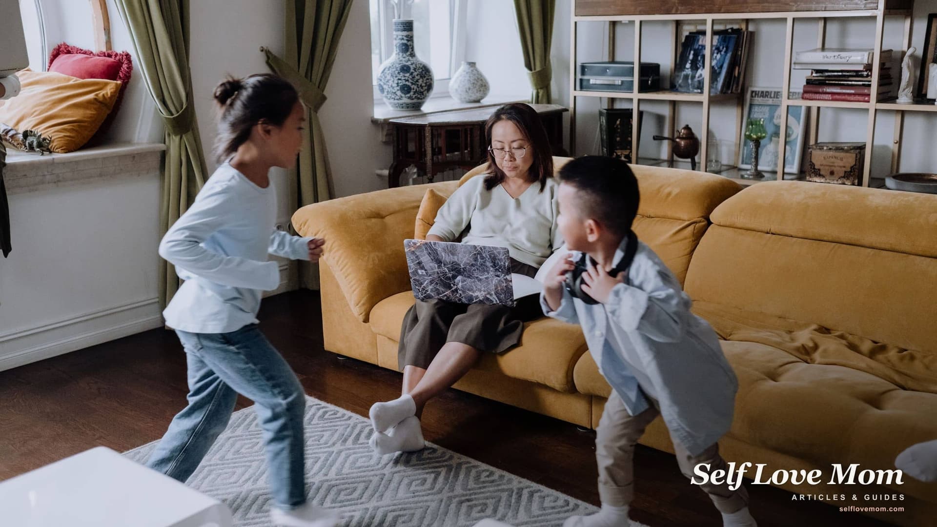 A mother checks housing programs on her laptop on the sofa while her two kids play around her in the living room.
