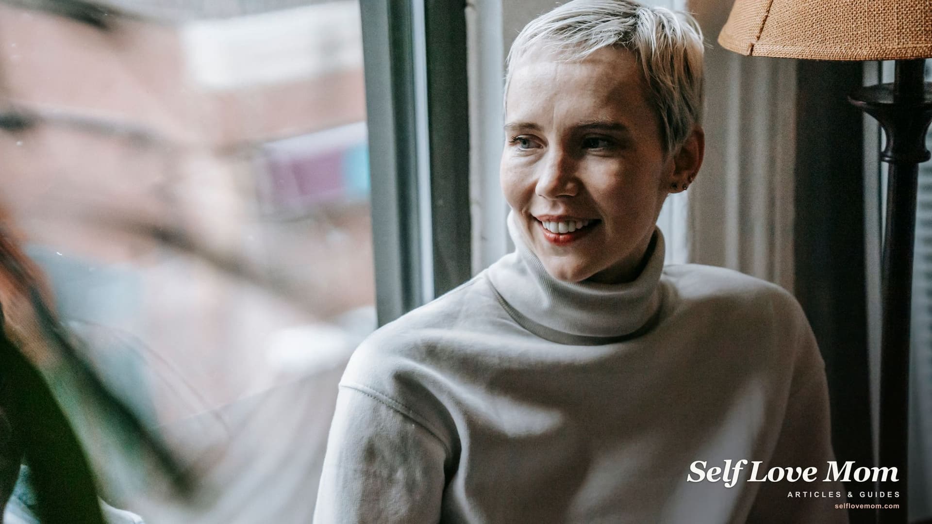 A smiling woman with short blond hair, in a white turtleneck, looks out of a bright window, at ease and sure of herself, showing how to feel confident again after divorce.