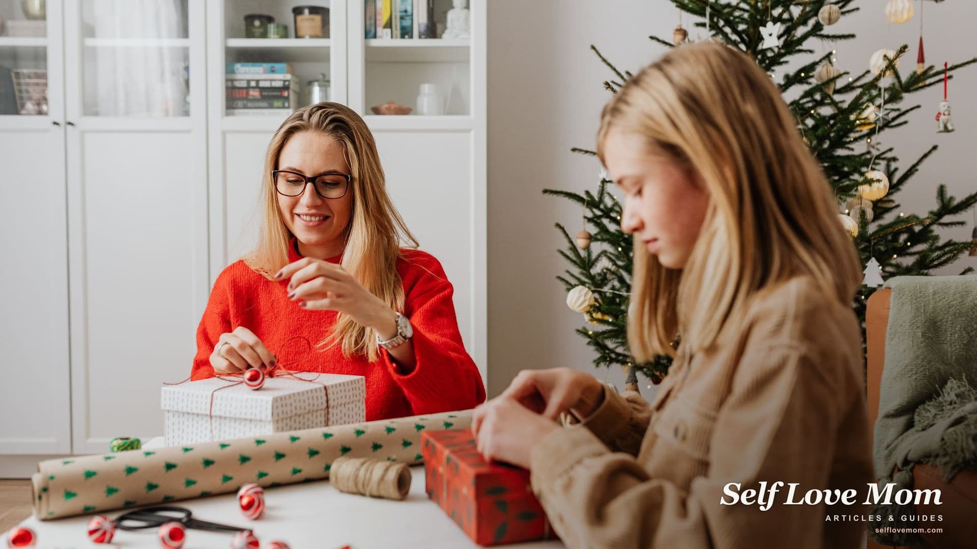 A mother in a red sweater wraps holiday gifts at a table beside the Christmas tree while her daughter ties ribbon on a present.