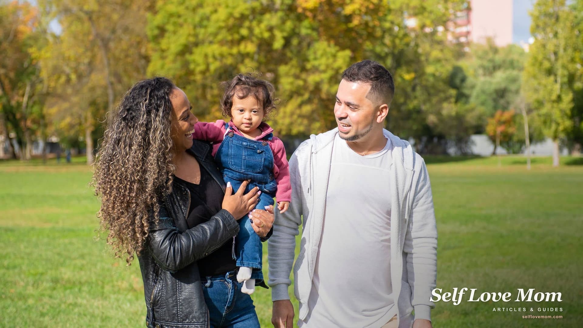 A mother holds her toddler daughter and smiles at a man walking beside them in a sunny park, the kind of short, public first meeting experts suggest.