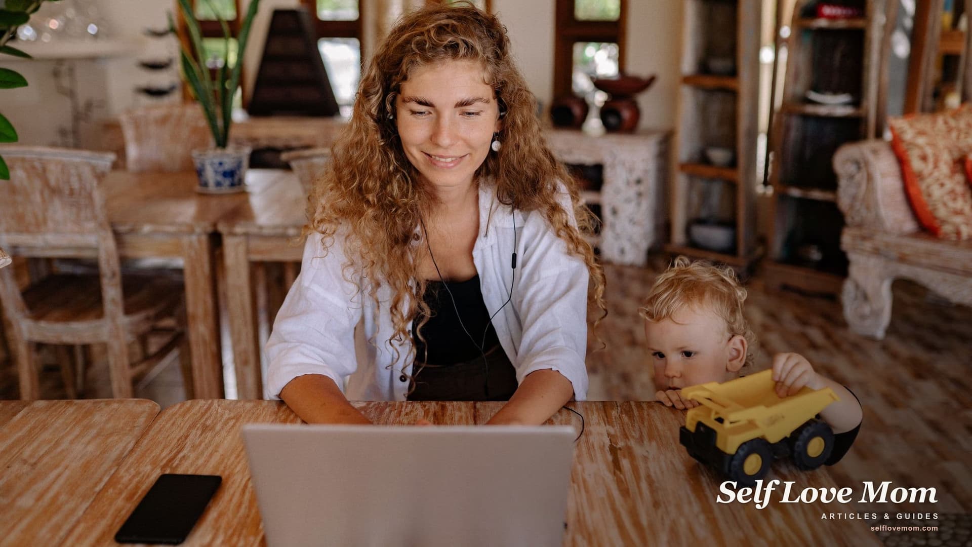 A smiling mom with earbuds works on her laptop at a wooden table while her toddler peeks over the edge with a toy truck, the kind of flexible setup many single mothers look for.