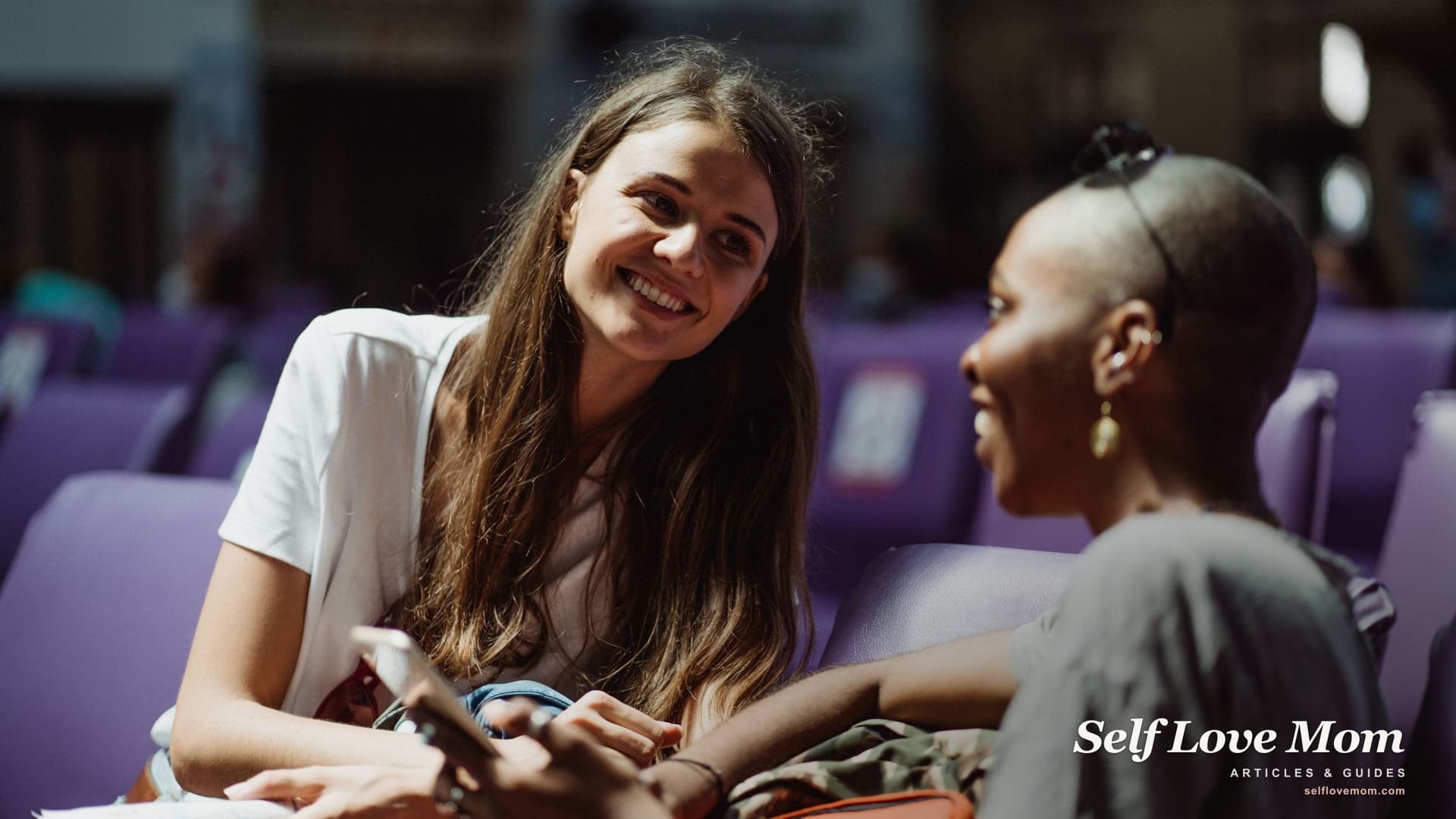 Two women smile and talk side by side while one holds her phone, the kind of easy first meetup many moms hope to find.