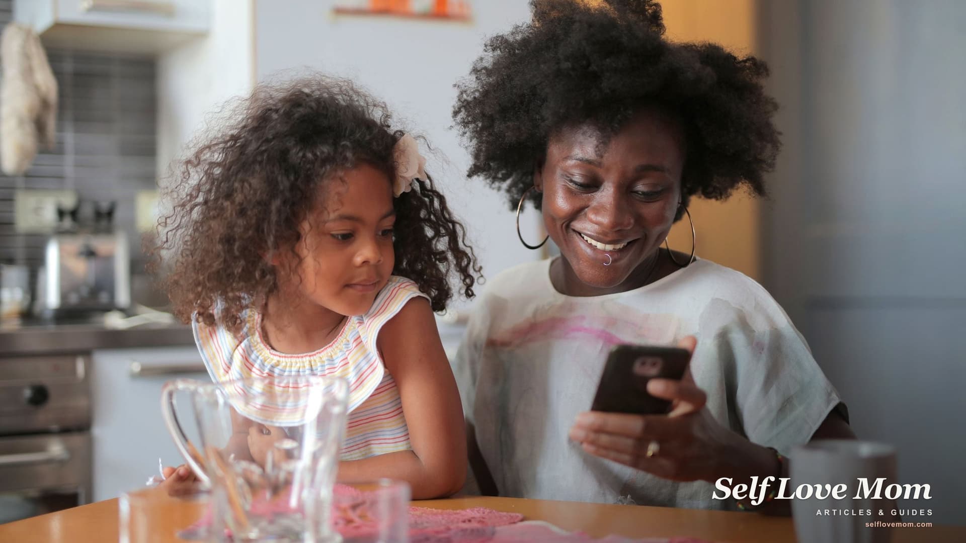 A smiling single mother reads a message on her phone at the kitchen table while her young daughter leans in beside her, the everyday side of using OurFamilyWizard.