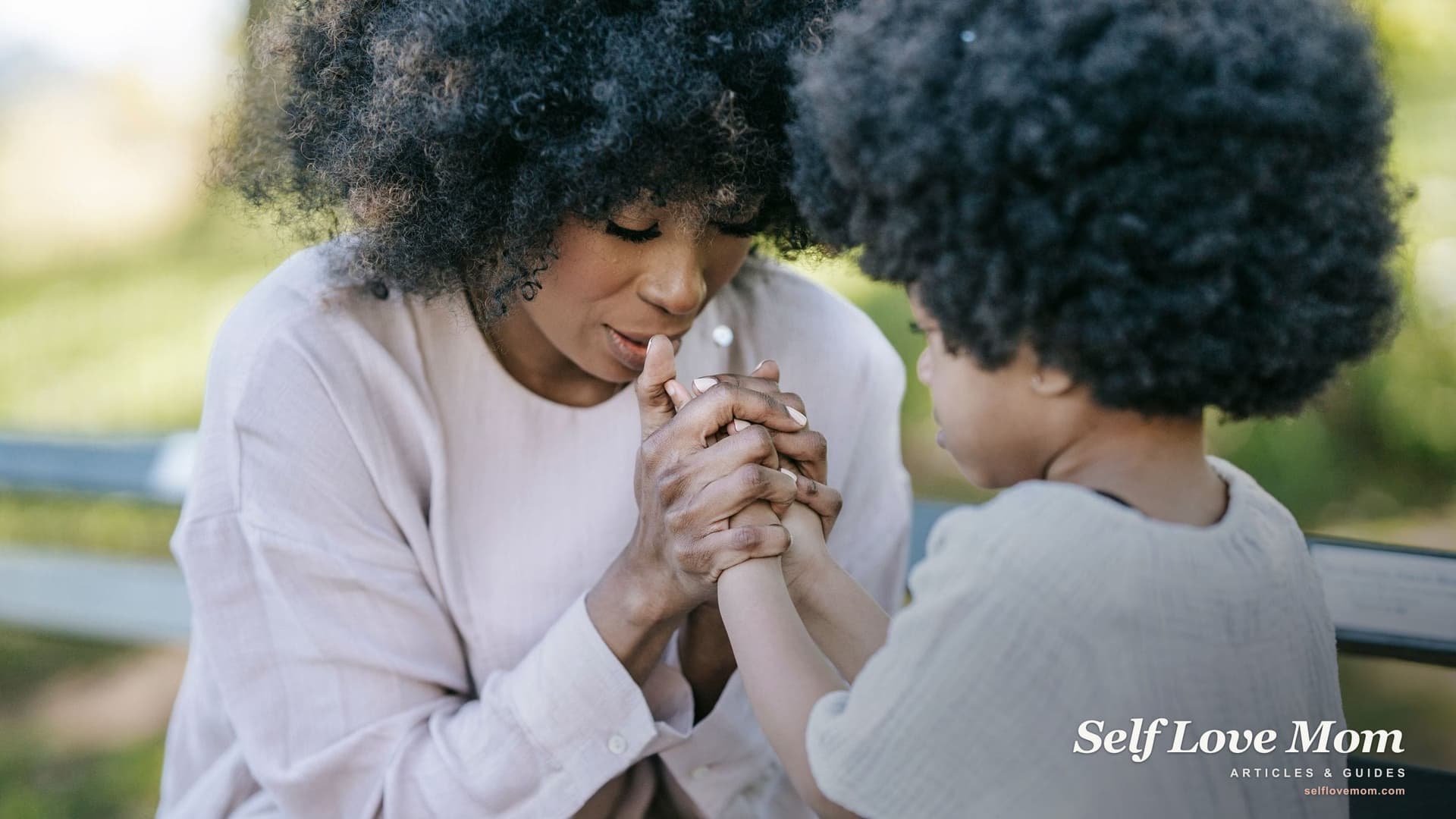 A mother sits close to her young child outdoors, holding the child's hands and speaking softly to comfort her.