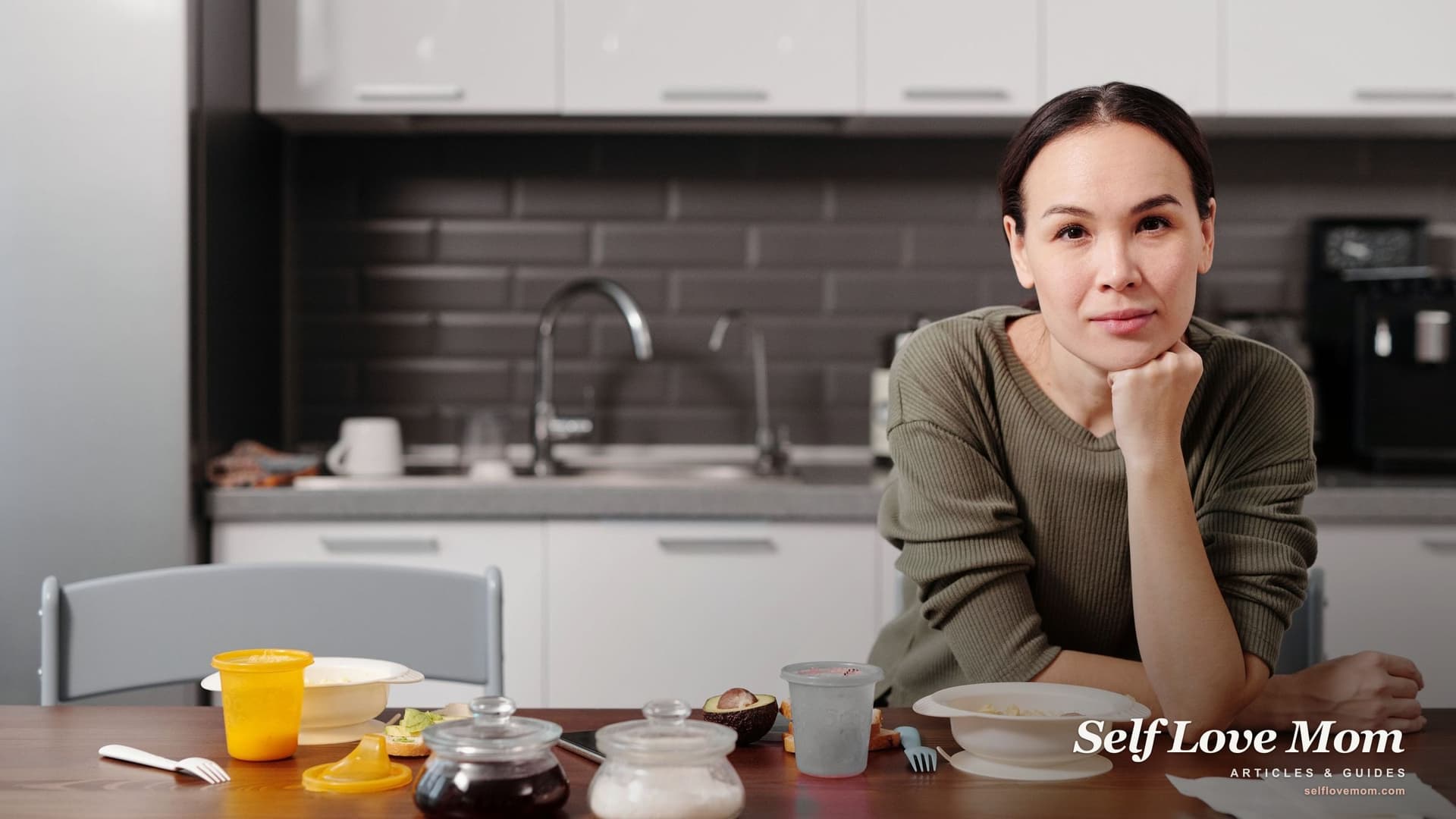 A mother sits at the kitchen table among her toddler's cups and bowls, chin on her hand, thinking over the signs your husband wants a divorce.