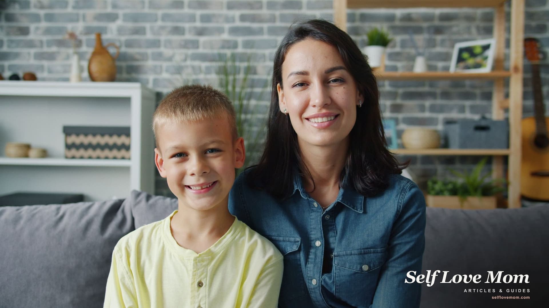 A smiling single mother sits on the couch at home next to her young son, both looking at the camera