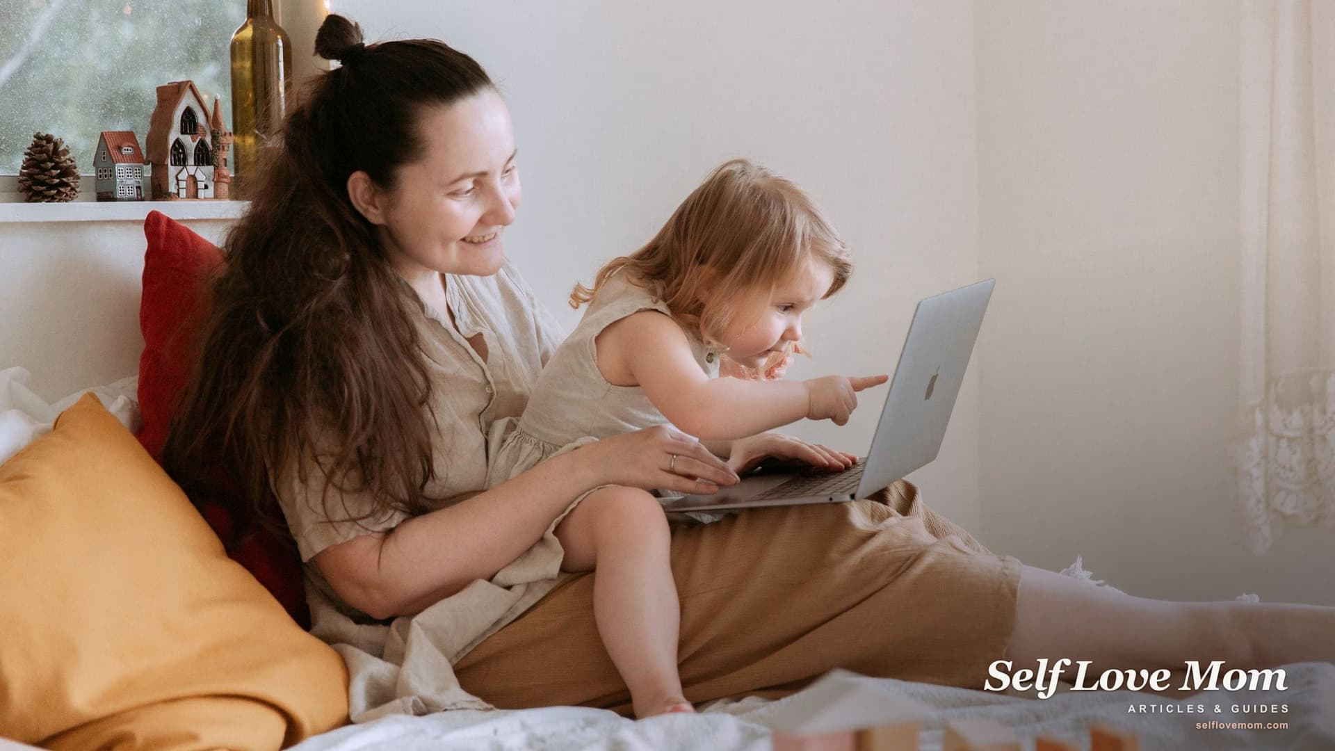 A mother smiles at her toddler, who points at the laptop screen on her lap, a work-from-home setup many virtual assistant jobs for moms involve.