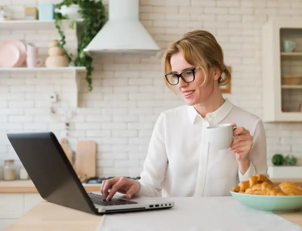 A working mom enjoying her coffee and working on her laptop in the kitchen. The image emphasizes self care for working moms by balancing work and personal moments