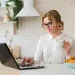 A working mom enjoying her coffee and working on her laptop in the kitchen. The image emphasizes self care for working moms by balancing work and personal moments