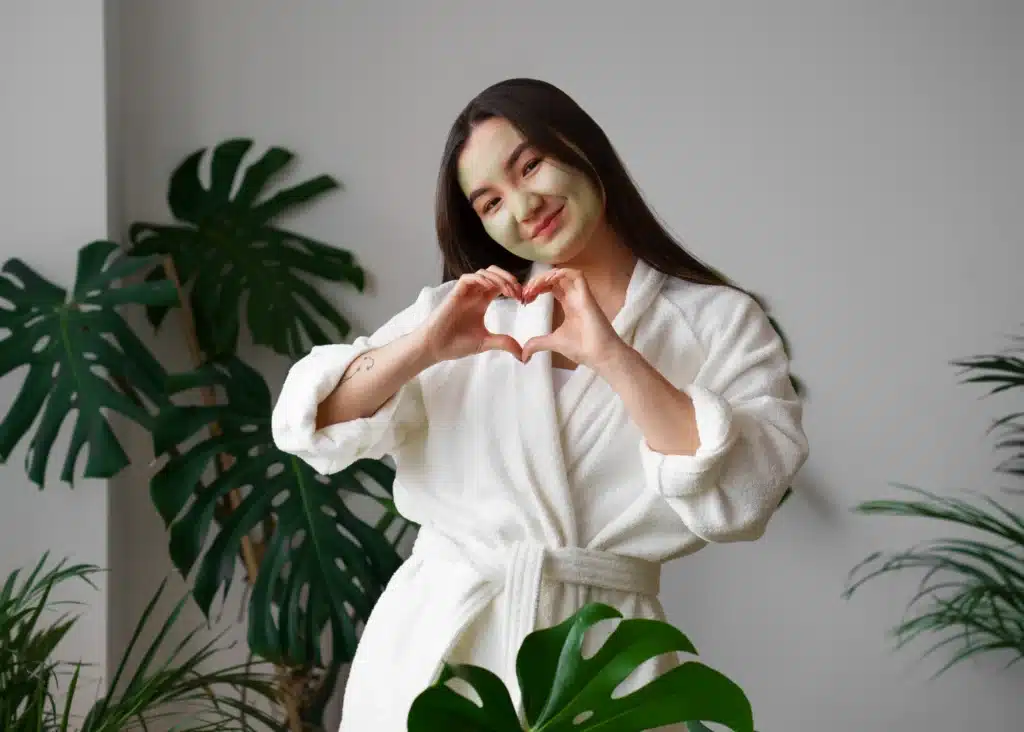 Woman in a white robe with a face mask making a heart shape while enjoying self-care at home, one of the simple things to do for yourself.
