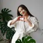 Woman in a white robe with a face mask making a heart shape while enjoying self-care at home, one of the simple things to do for yourself.