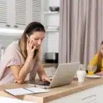 A mom working from home while talking on the phone and typing on her laptop in the kitchen, with her child sitting nearby. This setup illustrates the flexibility of jobs for mom at home.