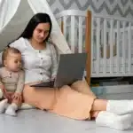 A stay-at-home mom working on a laptop while caring for her baby at home, representing flexible remote jobs for stay at home moms.