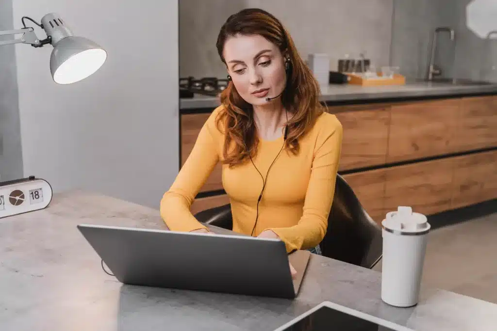 A woman working from home, wearing a yellow shirt and headphones, focused on her laptop. She’s engaged in a creative or content job, such as a voice-over artist, with a coffee cup and tablet nearby in a modern kitchen setting.